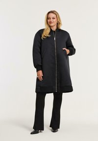Vrouw in een lange zwarte gewatteerde jas met ritssluiting, zwarte flared broek en zwarte puntige hakken, staand tegen een effen achtergrond.