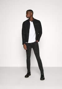 Zwarte suède jas met ritszak, witte shirt, zwarte skinny jeans en zwarte enkellaarzen, gestyled tegen een witte achtergrond.