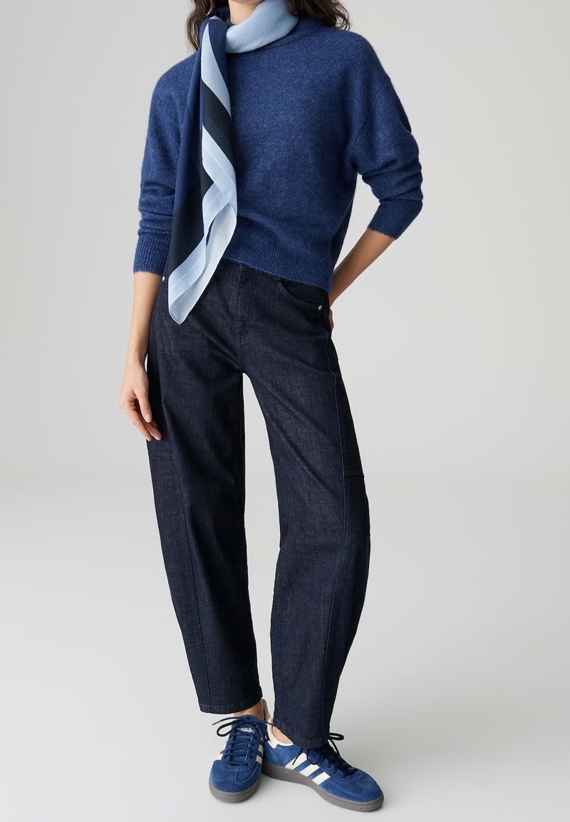 Femme portant un pull bleu, un jean large foncé, une écharpe bleue et blanche, et des baskets bleues, debout avec une main sur la hanche.
