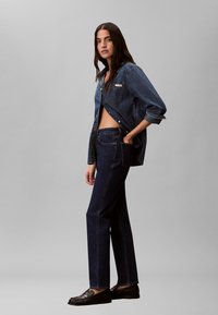 Ženska stoji postrani, oblečena v temno denim srajco z zavihanimi rokavi, usklajene jeans hlače in črne loafers čevlje, na enobarvni sivi podlagi.