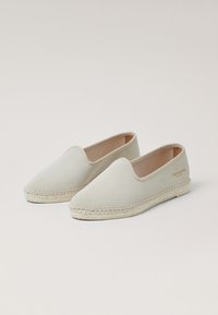 Beige Leinenschlüpfschuhe mit einer strukturierten Oberfläche, ausgestattet mit einer gewebten Jutesohle und minimalistischem Design. Dies beinhaltet dezentes Branding an der Seite.