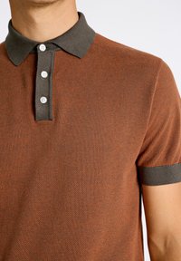 Kortärmad polo i rostfärg med mörkgrå krage och muddar. Har en texturerad tyg och tre-knapps knäppning.