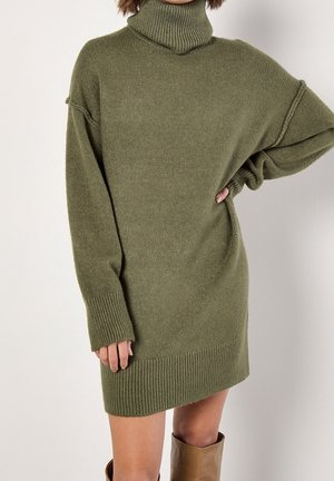 Femme portant une robe-pull à col roulé côtelé vert olive avec manches longues et bottes marron montantes jusqu'aux genoux.