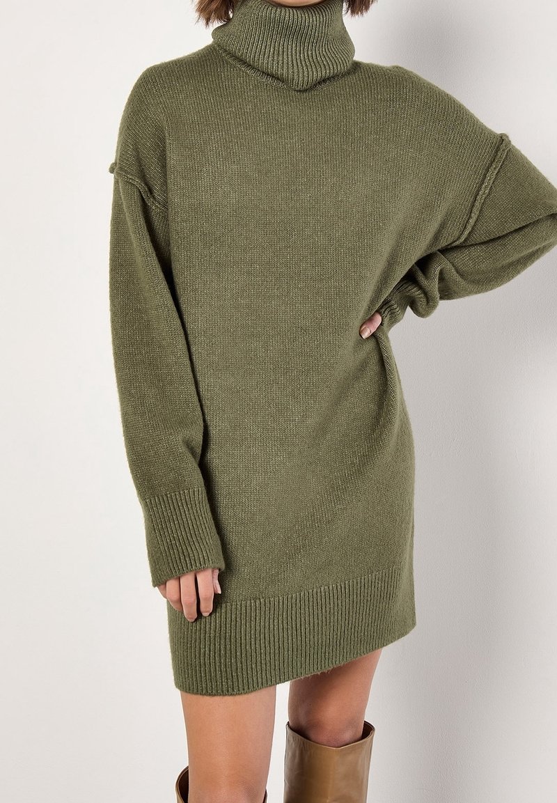 Femme portant une robe-pull à col roulé côtelé vert olive avec manches longues et bottes marron montantes jusqu'aux genoux.