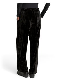 Personne portant un pantalon ample en velours noir et des chaussures noires, debout avec une main dans la poche, vue de dos sur fond blanc.