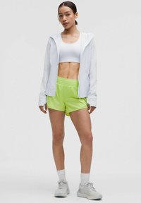 White lightweight zip-up jacket, fitted white crop top, and bright green shorts. Model wears light gray athletic shoes and white socks.