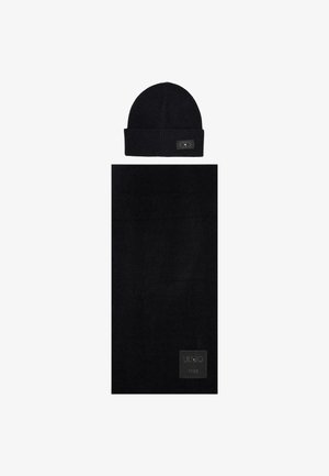 Chapeau en tricot noir avec un revers retourné et une petite étiquette, associé à une écharpe noire assortie présentant un motif texturé et un patch logo.