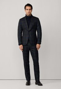 Uomo che indossa un completo a quadri verde scuro e blu navy con pantaloni coordinati, maglione a collo alto nero e scarpe eleganti in pelle nera.