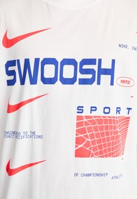 Balta kokvilnas T-kreklā ar izteiksmīgu zilu "SWOOSH" tekstu, sarkaniem Nike logo akcentiem un sarkanu režģa rakstu kopā ar teksta blokiem.