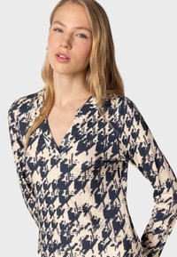 Un haut à manches longues et en V, fabriqué en tissu doux, avec un motif abstrait bleu marine et beige. Le design présente une silhouette ajustée et une texture lisse.