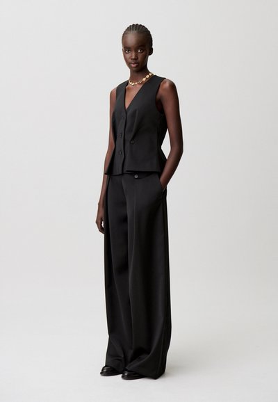 Chaleco sin mangas negro con escote en V, dos botones y bolsillos delanteros, combinado con pantalones de pierna ancha en tela y color a juego.