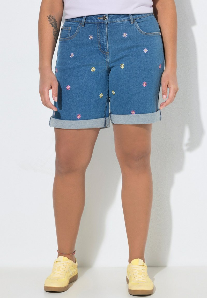 Pantalones cortos de mezclilla con dobladillos enrollados y flores coloridas bordadas, combinados con zapatillas amarillas y camiseta blanca, mostrando piernas y brazo tatuado.