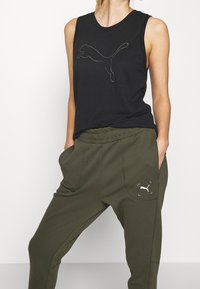 Schwarzes ärmelloses Top mit einem springenden Puma-Logo, kombiniert mit olivgrünen Sweatpants mit einem weißen Logo auf der Tasche. Weicher Stoff, lockere Passform.
