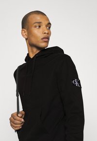 Čierna mikina s kapucňou z mäkkej tkaniny, s klokankou, nastaviteľnou kapucňou na šnúrku a značkovou nášivkou Calvin Klein na rukáve.