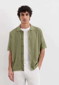 Short-sleeve, collared shirt in light olive green fabric, featuring a textured surface and button closure, worn over a white t-shirt.
