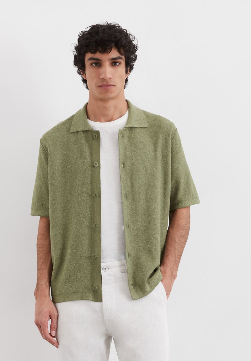 Short-sleeve, collared shirt in light olive green fabric, featuring a textured surface and button closure, worn over a white t-shirt.