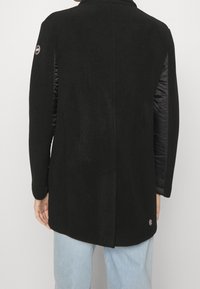 Manteau noir avec un corps en laine, des manches en nylon matelassé et un accent de logo sur le bas à gauche. Silhouette droite, légèrement plus longue à l'arrière.