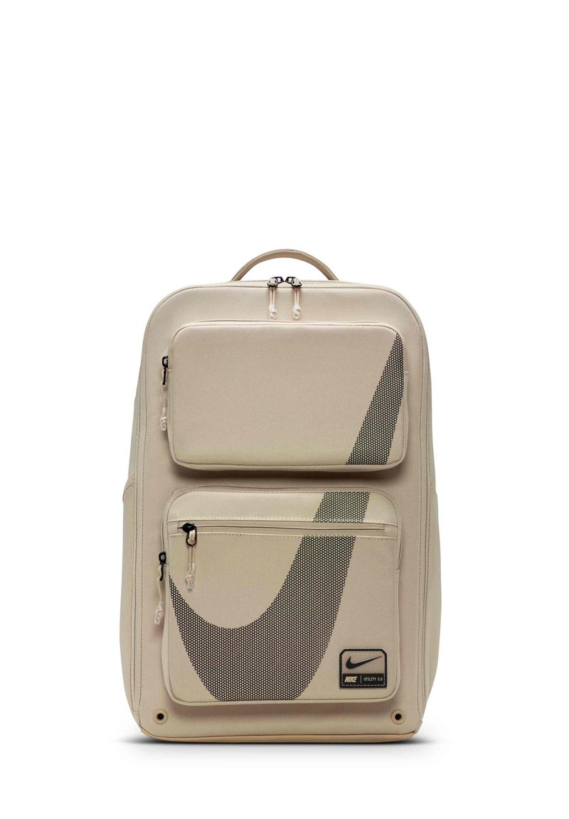 Beige rygsæk med mesh-paneler, dobbelte lynlåslommer, sort Nike-logo og robust håndtag. Glat stof med geometriske designelementer.