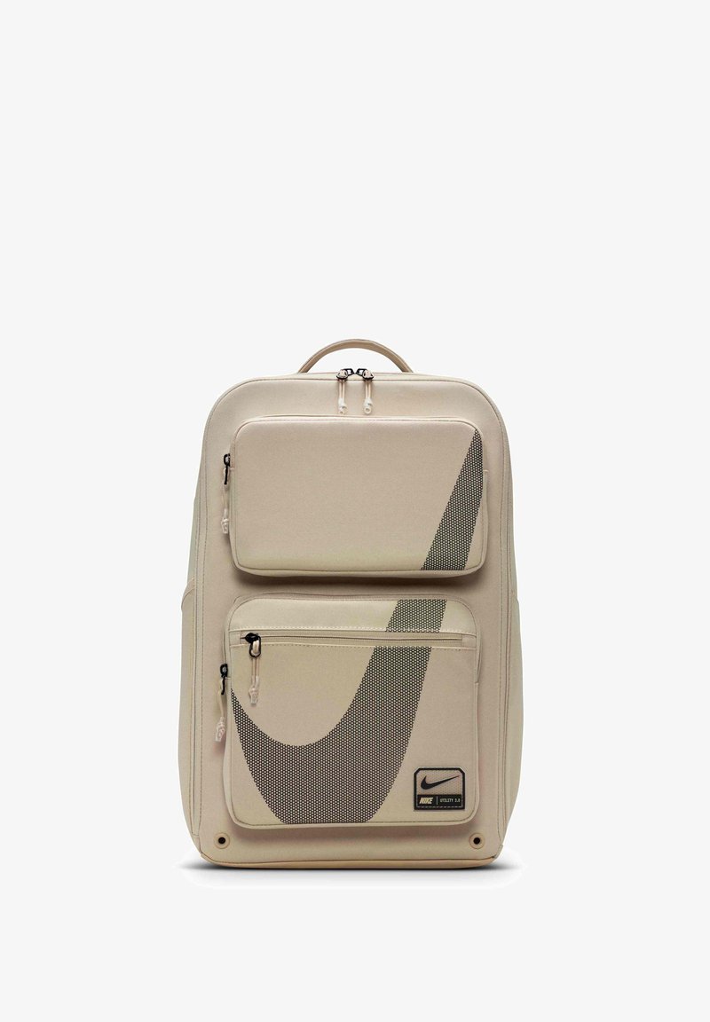 Beige rygsæk med mesh-paneler, dobbelte lynlåslommer, sort Nike-logo og robust håndtag. Glat stof med geometriske designelementer.