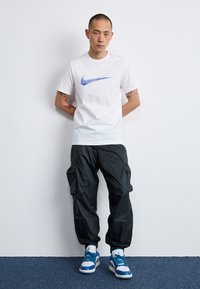 White t-shirt with blue Nike logo, black cargo pants with pockets, and blue and white sneakers with laces. Stands against a plain wall.