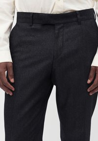 Homme portant un pantalon noir texturé et une chemise crème à manches longues, mains reposant près des côtés, concentré sur la taille et le haut des jambes.