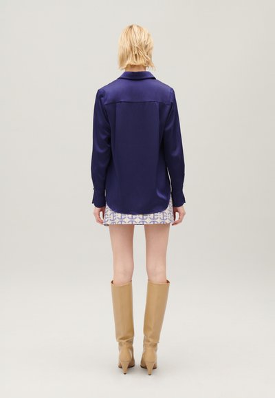 Persona bionda con capelli corti in piedi di spalle, indossa una blusa a maniche lunghe blu scuro, una gonna con fantasia e stivali al ginocchio color beige.