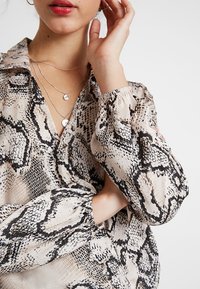 Donna che indossa una blouse beige e nera con stampa a serpente, maniche a sbuffo e delicate collane di argento a più strati, mano vicino al viso.