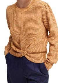 Pull tricoté jaune moutarde avec une texture côtelée. Il présente des manches longues et un détail torsadé à l'ourlet avant, assorti à un pantalon bleu marine.