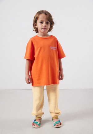 Bambino in piedi, indossa una maglietta arancione oversize con la scritta "American Vintage", pantaloni crema e sandali multicolore su uno sfondo semplice.
