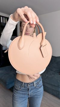 Στρογγυλή ροζ-πορτοκαλί δερμάτινη τσάντα με διπλές λαβές από πάνω, λεία υφή και μια μικρή προσαρτημένη ετικέτα. Απλός, μινιμαλιστικός σχεδιασμός.