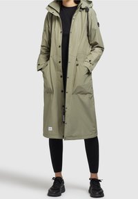 Long imperméable kaki avec une capuche, des boutons à l'avant, de grandes poches et un cordon de serrage ajustable. Porté sur un haut noir et des leggings.