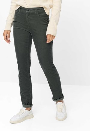 Personne portant un pantalon slim vert foncé, des baskets blanches et un pull en maille couleur crème, debout devant un fond blanc uni.
