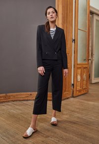 Black cropped double-breasted blazer with two buttons, worn over a black and white striped top. Black wide-leg trousers and white slide sandals.