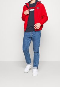 Rød hættetrøje med lynlås over en marineblå sweater med hvide og røde striber, parret med lyseblå jeans og hvide sneakers.
