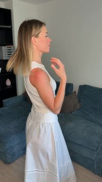 Jeune femme aux cheveux blonds mi-longs portant une robe blanche, debout dans un salon avec un canapé bleu.
