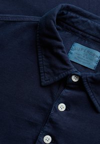 Chemise bleu marine avec col, boutons blancs et une étiquette de marque bleue visible à l'intérieur du col.