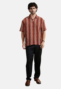 Camisa de manga corta de color rojizo con un patrón de formas geométricas, combinada con pantalones negros y zapatos de cuero marrón sin cordones.