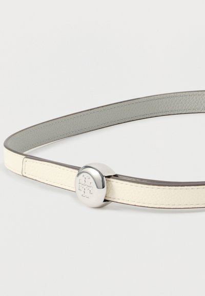 Ohut kermaisten nahkavyön, jossa on harmaa alapuoli ja pyöreä hope buckle, joka on kaiverrettu logolla, esitelty valkoista taustaa vasten.