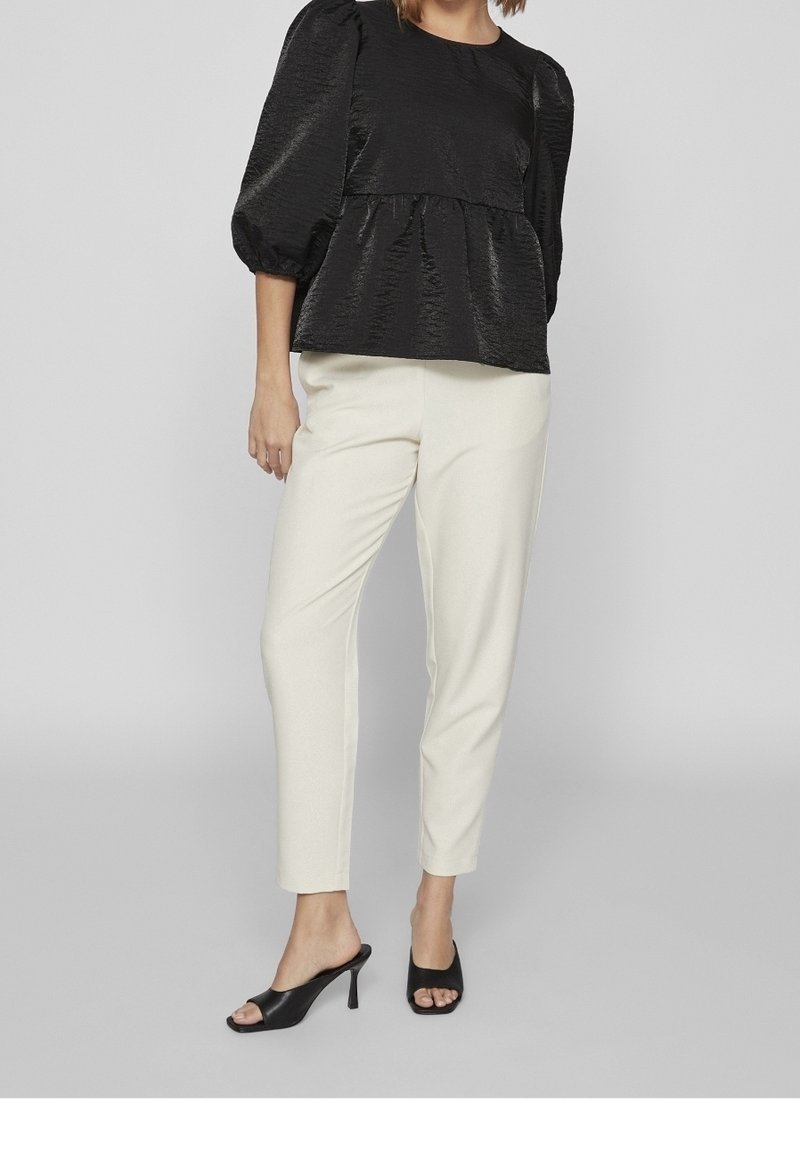 Personne portant un chemisier noir à manches bouffantes, un pantalon blanc fuselé et des mules noires à talons ouvertes, debout devant un fond uni.