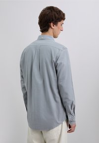 Lysegrå langærmet skjorte lavet af blødt stof. Har afrundet kant, knaplukning ved ærmerne og en lille læg bagpå for ekstra komfort.