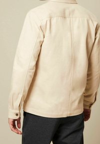 Giacca in tela beige chiaro con colletto appuntito, polsini abbottonati e dettagli sul retro, mostrata da un'angolazione posteriore.