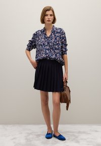 Navyfarbene Bluse mit Paisley-Muster und umgeschlagenen Ärmeln, kombiniert mit einem gefalteten schwarzen Rock. Das Model hält eine braune Tasche und trägt blaue Ballerinas.