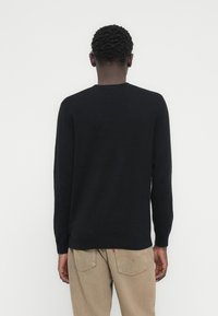 Pull en maille noire avec un col rond, manches longues et poignets côtelés, porté avec un pantalon beige, vu de dos sur un fond uni.