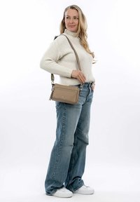 Bolso cruzado de cuero beige con un bolsillo con cremallera, herrajes plateados y una correa ajustable, combinado con jeans azul claro y zapatillas blancas.