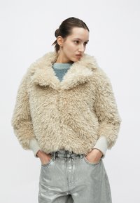 Beiger Kunstpelzjacke mit weitem Kragen, flauschiger Textur und gerippten Bündchen. Getragen über einem hellblauen Pullover und metallisch silbernen Hosen.