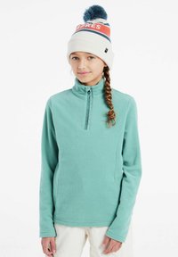 Felpa in pile di un azzurro chiaro con colletto alto e zip a metà, caratterizzata da una texture morbida. Abbinata a un berretto lavorato a maglia bianco con un pom-pom blu.