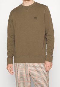 Felpa marrone con colletto a girocollo, caratterizzata da un logo a patch testurizzato sul petto e cuciture a contrasto. Indossata con pantaloni a scacchi.