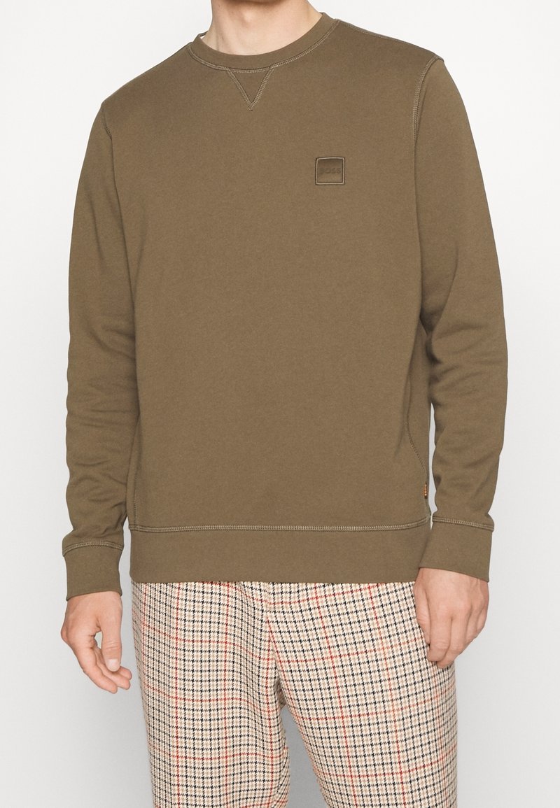 Felpa marrone con colletto a girocollo, caratterizzata da un logo a patch testurizzato sul petto e cuciture a contrasto. Indossata con pantaloni a scacchi.