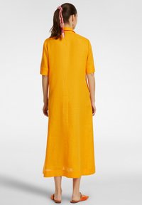 Lunga vestito arancione brillante in tessuto leggero. Presenta maniche corte e un colletto, con un taglio dritto che arriva a metà polpaccio.