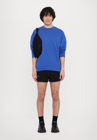 Felpa blu, pantaloni shorts neri e una borsa a tracolla nera. Il modello è in piedi con capelli ricci corti e indossa scarpe e calze nere.